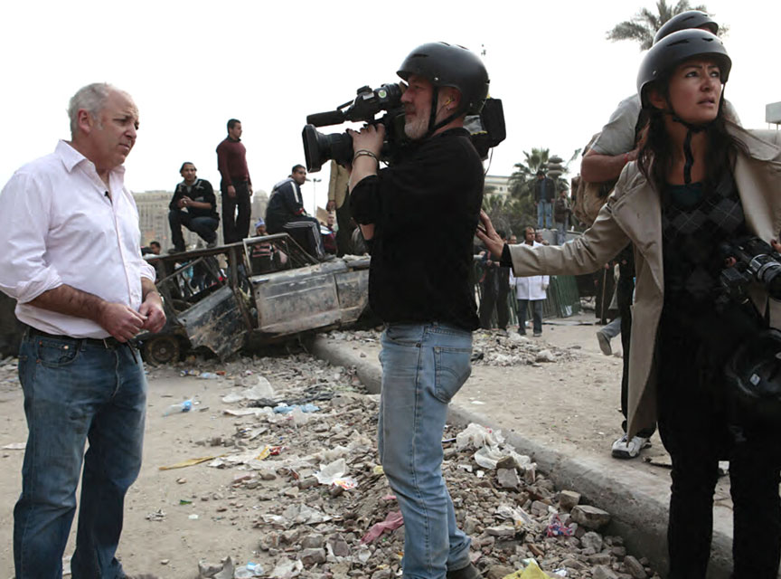 Jeremy Bowen in front of camera with nervous producer in war zone